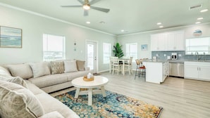 A spacious living room with a sofa, coffee table, and a ceiling fan. There is a dining area with a table and chairs, and a kitchen with white cabinets and a sink. - Spacious 3 Bedroom Surfside Home Near The Jetties (Surfside Beach)