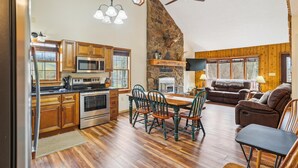 A spacious kitchen with wooden cabinets, a dining area with a wooden table and chairs, a stone fireplace, and a cozy living area with a sofa and a television. - Come stay! Sam's Cabin is a genuine mountain treasure. (Canaan Valley)