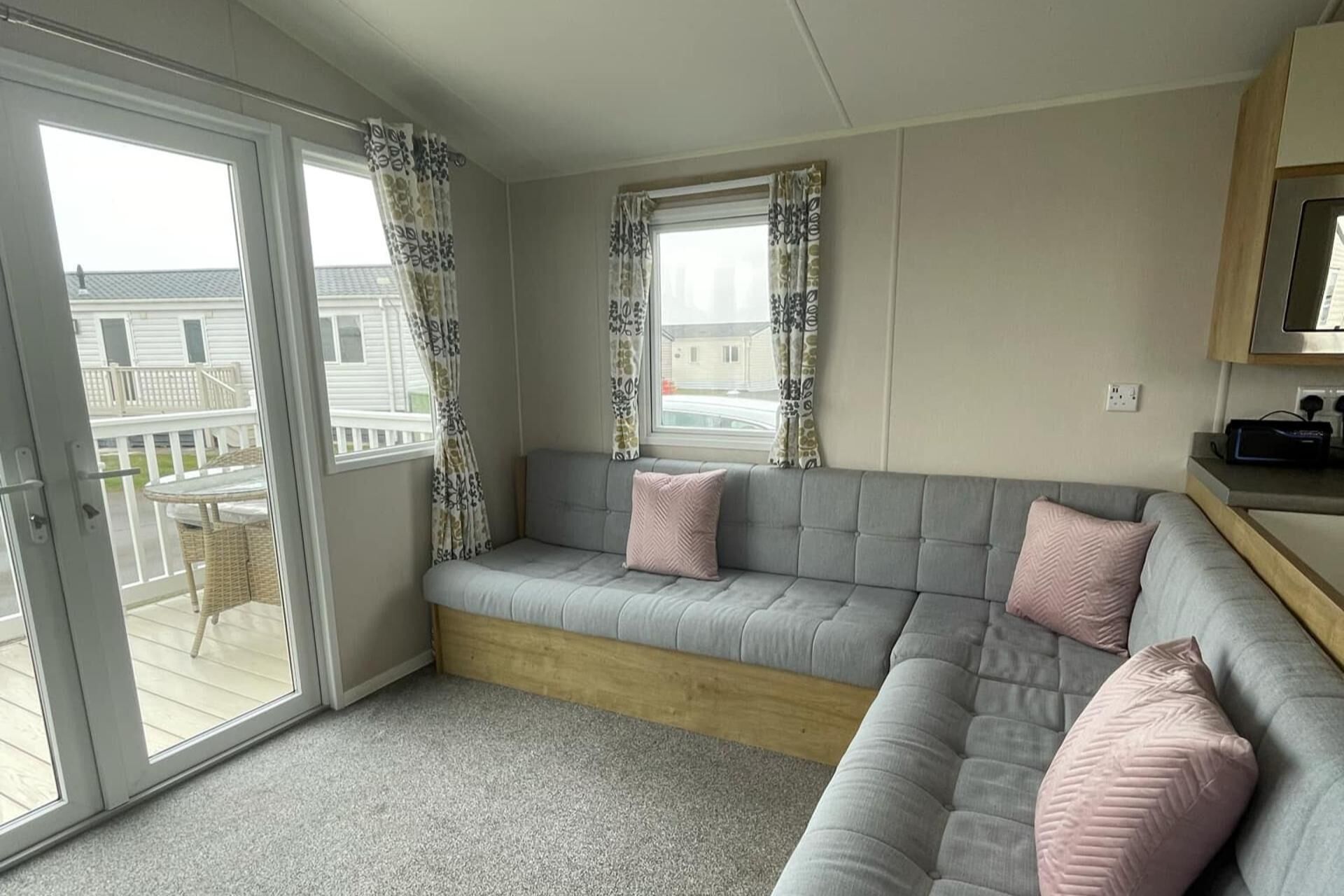 A modern living room with a grey sofa, two pink cushions, a wooden bench, and a sliding glass door leading to an outdoor balcony.