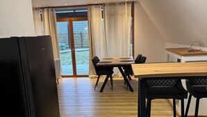 Une cuisine compacte avec un réfrigérateur noir, une table à manger en bois et quatre chaises, et une vue sur un jardin grâce à de grandes fenêtres.