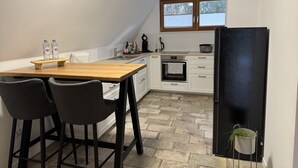 Une cuisine de taille réduite, avec une table en bois, deux tabourets de bar, un réfrigérateur noir et une fenêtre avec des stores.