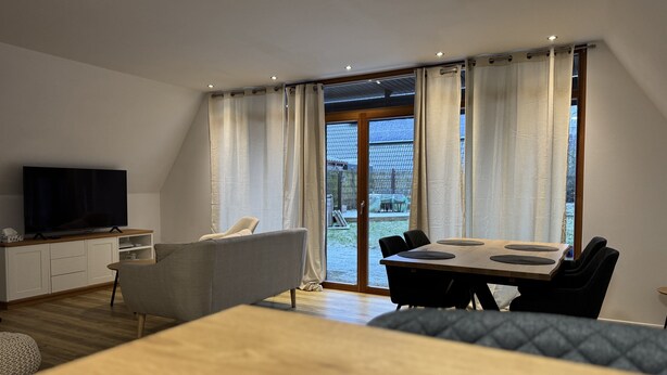 Un salon moderne avec un téléviseur à écran plat, un canapé gris, une table à manger en bois avec des chaises noires et de grandes fenêtres avec des rideaux.