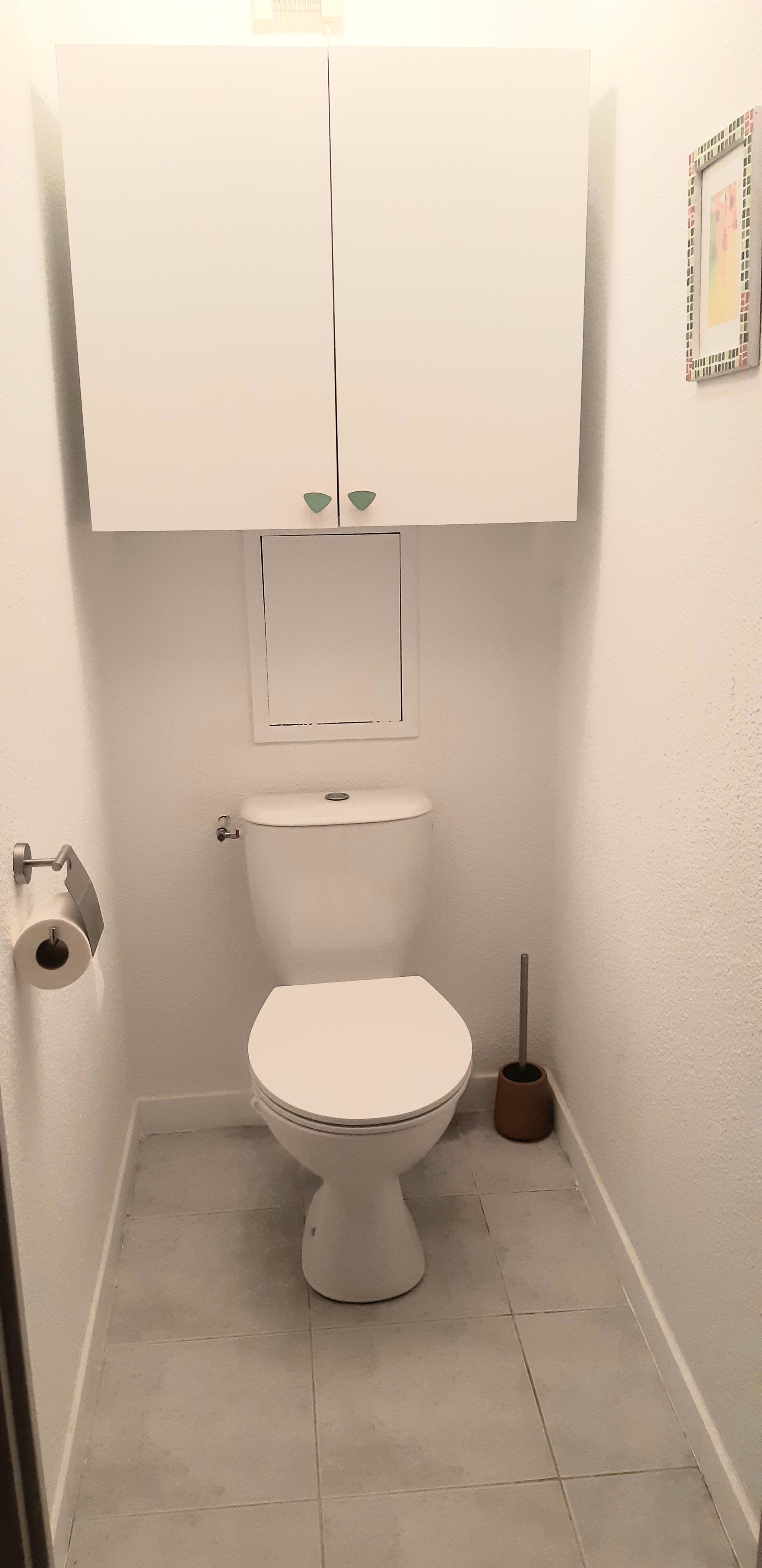 Un baño pequeño y blanco con inodoro, dispensador de papel higiénico y escobilla.
