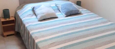 Habitación individual con ropa de cama a rayas, una mesita de noche con lámpara y una ventana con vista a la vegetación.