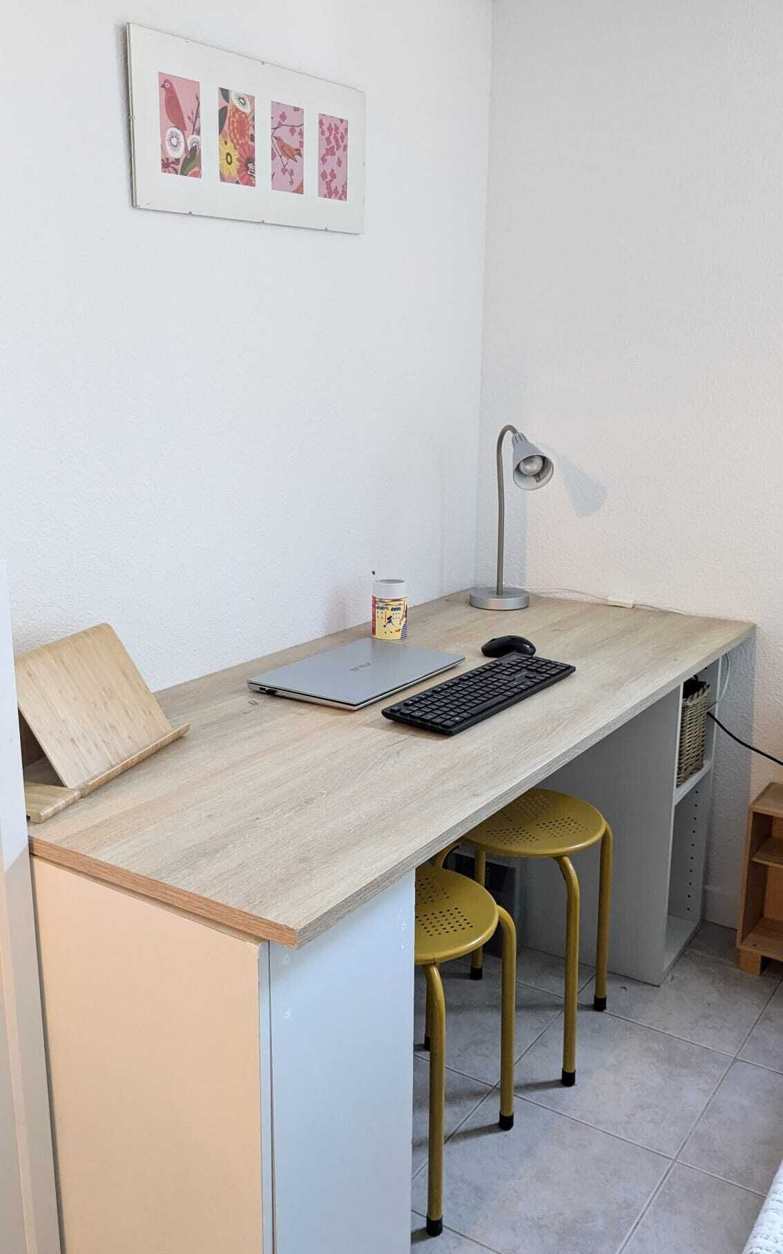 Un espacio de trabajo pequeño con una computadora portátil, teclado y lámpara de escritorio. Hay dos taburetes amarillos y un estante con una lata.