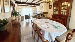 A dining area with a wooden table set for four, surrounded by chairs. A chandelier hangs from the ceiling, and a bookshelf filled with books is to the right. - La Baraque de Papi - Spacious detached house - Private garden (Marck)