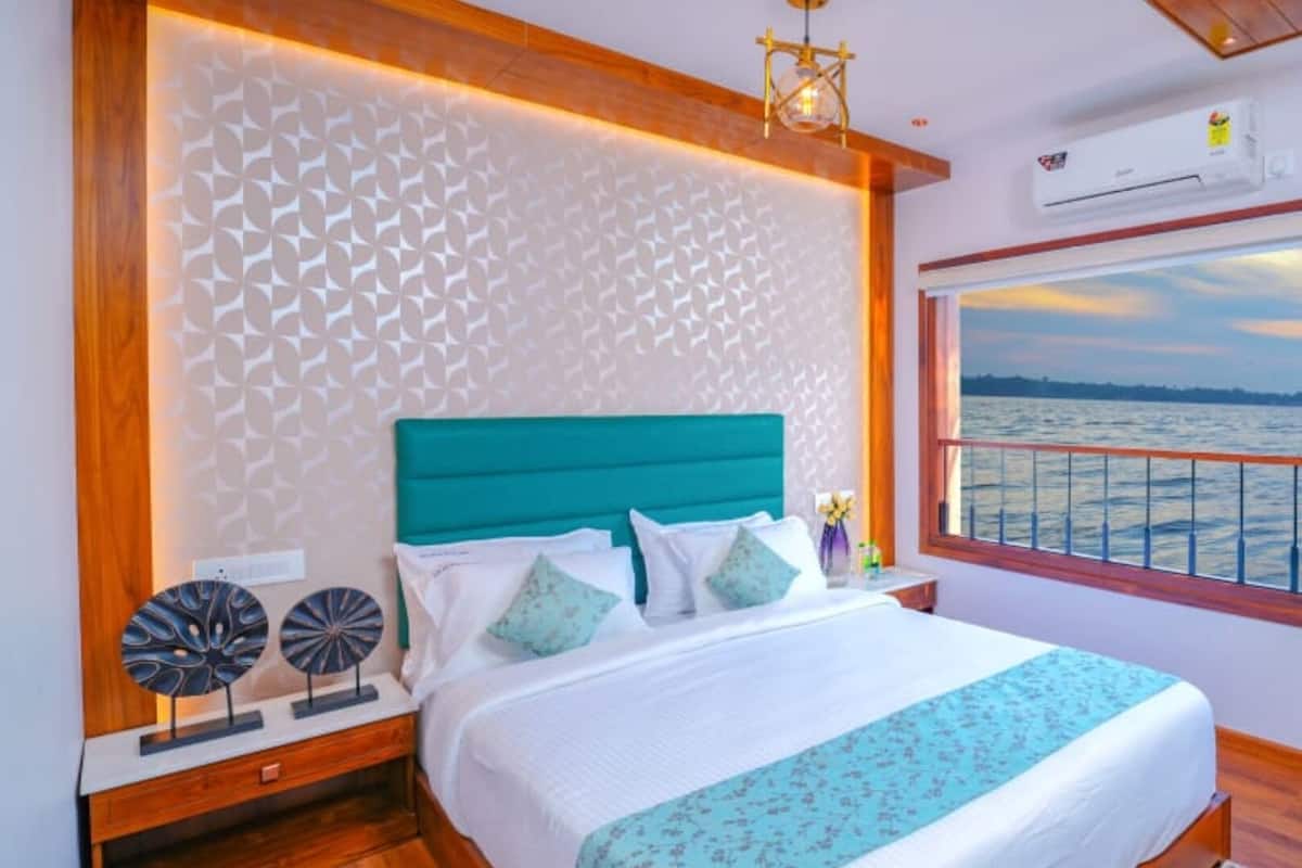 A bedroom with a large window offering a view of the water, a bed with white bedding and teal accents, and decorative wall art.