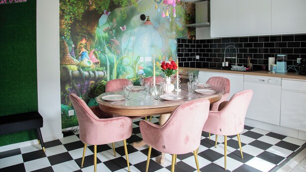 Zona pranzo con tavolo rotondo apparecchiato per sei, circondato da sedie rosa dalle gambe dorate. Al posto di una parete dietro il tavolo, un murale raffigura una scena boschiva fantastica.