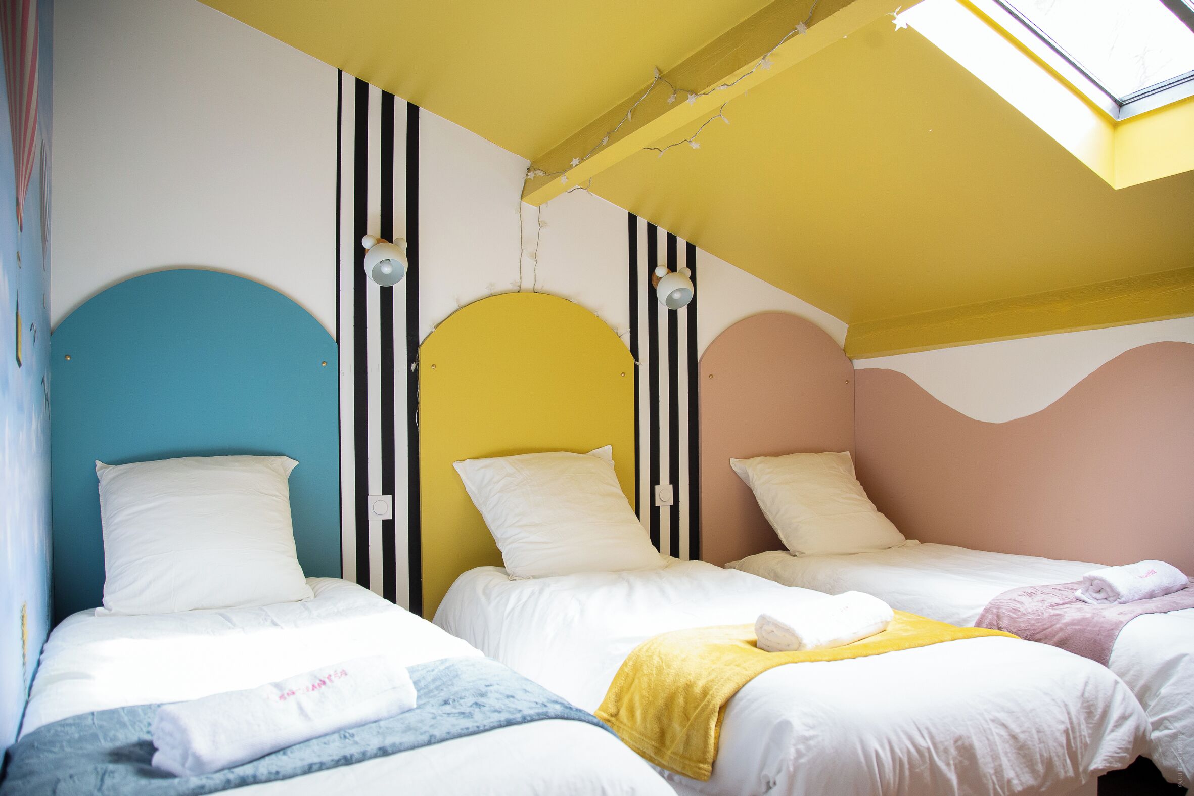 Een hotelkamer met drie bedden, elk met witte beddengoed en een uniek hoofdeind. De kamer heeft een schuine plafond met een dakraam en gestreepte wanden.