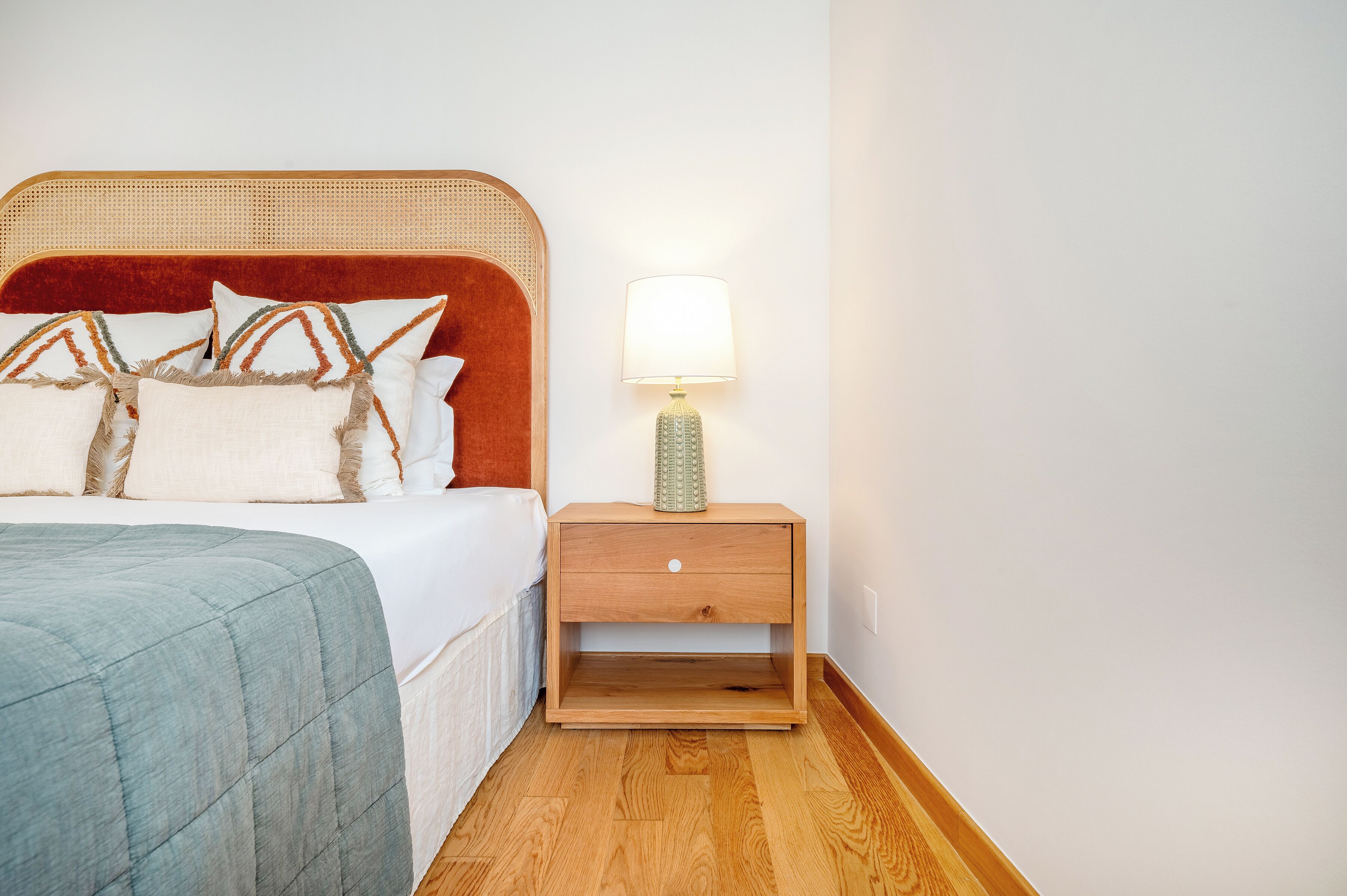 A neatly made bed with a red headboard, a bedside lamp, and a wooden nightstand with a drawer.