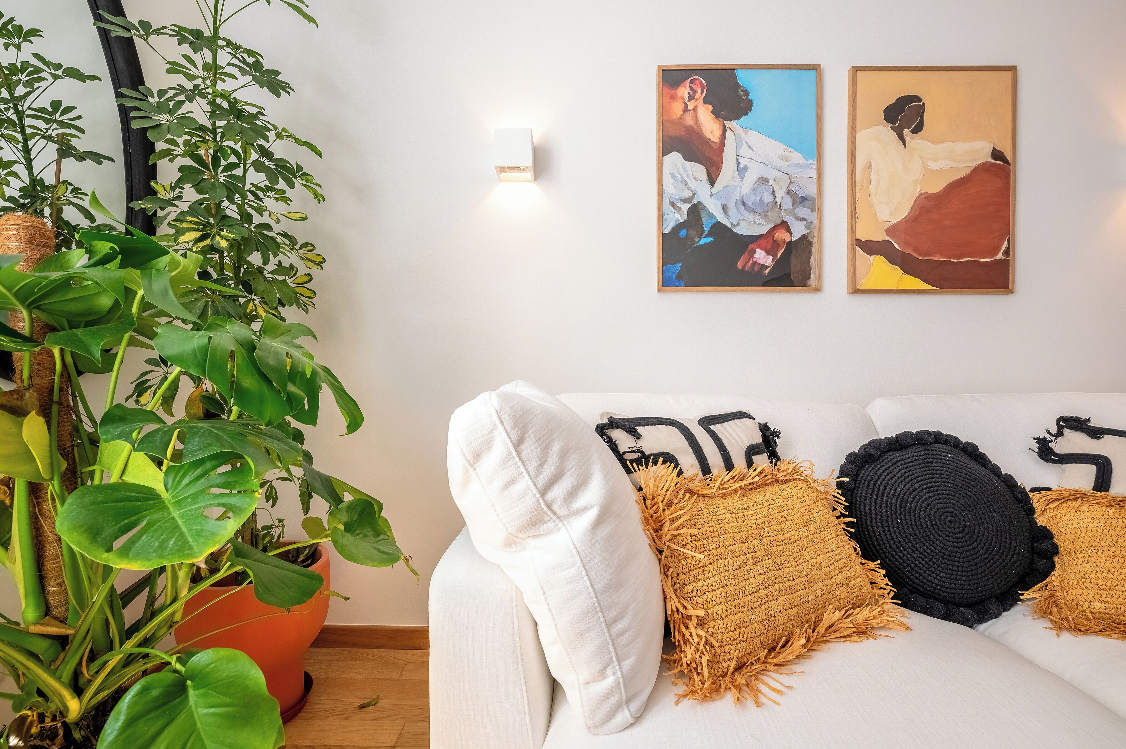 A living room with a white sofa, decorative pillows, a large green plant, and two framed artworks on the wall.