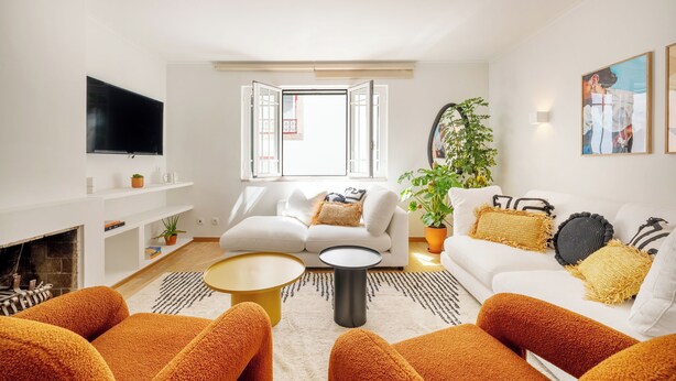 Una sala de estar moderna con un sofá blanco, sillones naranjas, un televisor de pantalla plana, una chimenea y un ventanal con contraventanas abiertas.