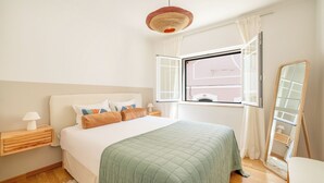 Un dormitorio con cama, mesita de noche, lámpara, ventana y espejo.