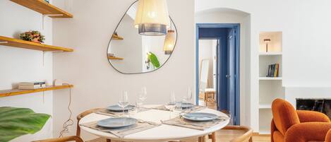 A dining area with a round table set for six, surrounded by wicker chairs. A large mirror hangs on the wall, reflecting the room.