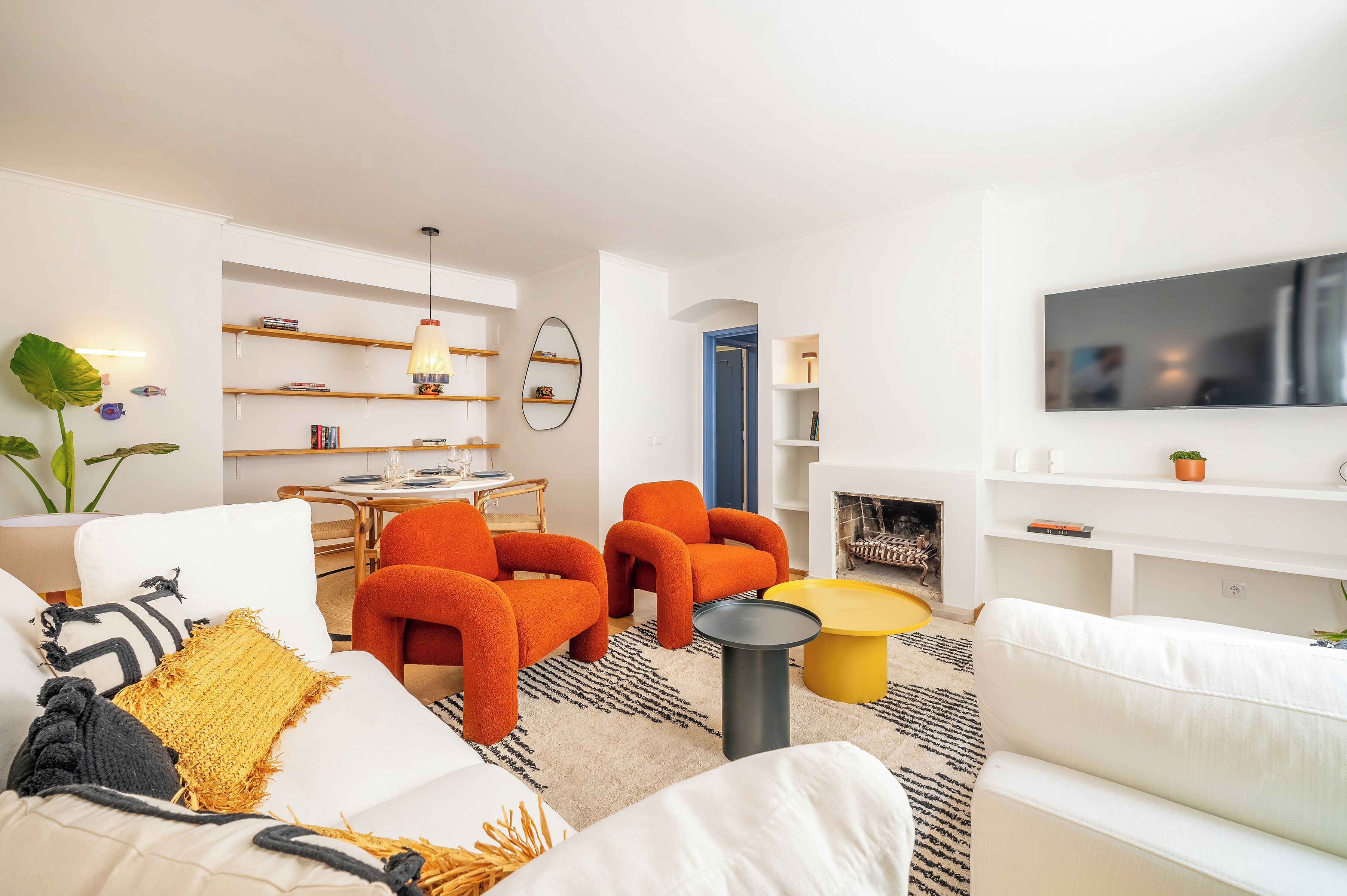 Uma sala de estar moderna com sofás brancos, poltronas laranja e uma mesa de centro amarela. Há prateleiras com livros e objetos decorativos, e uma TV de tela plana na parede.