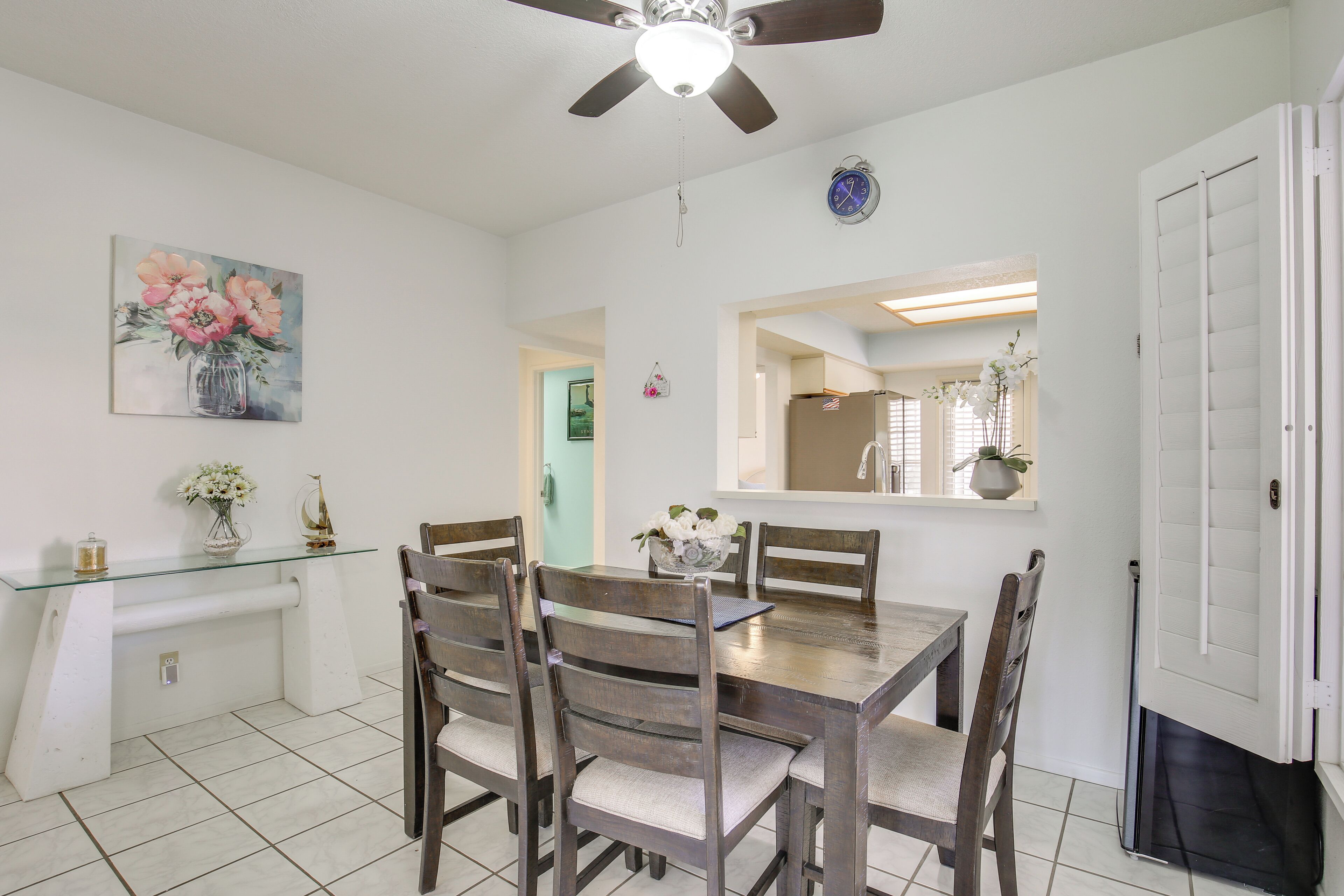 A dining area with a glass-top table, wooden chairs, a ceiling fan, and a painting on the wall.