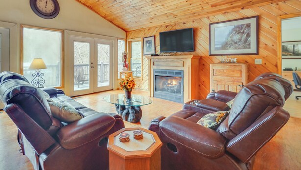 A cozy living room with leather sofas, a glass coffee table, a fireplace, and a television mounted on the wall.