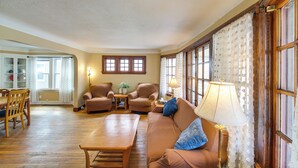 Sala de estar com piso de madeira, um sofá marrom, duas poltronas, uma mesa de centro e um abajur. Há janelas com cortinas e uma porta que dá para outro cômodo.