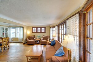 Una sala de estar con piso de madera, un sofá marrón, dos sillones, una mesa de centro y una lámpara. Hay ventanas con cortinas y una puerta que conduce a otra habitación.