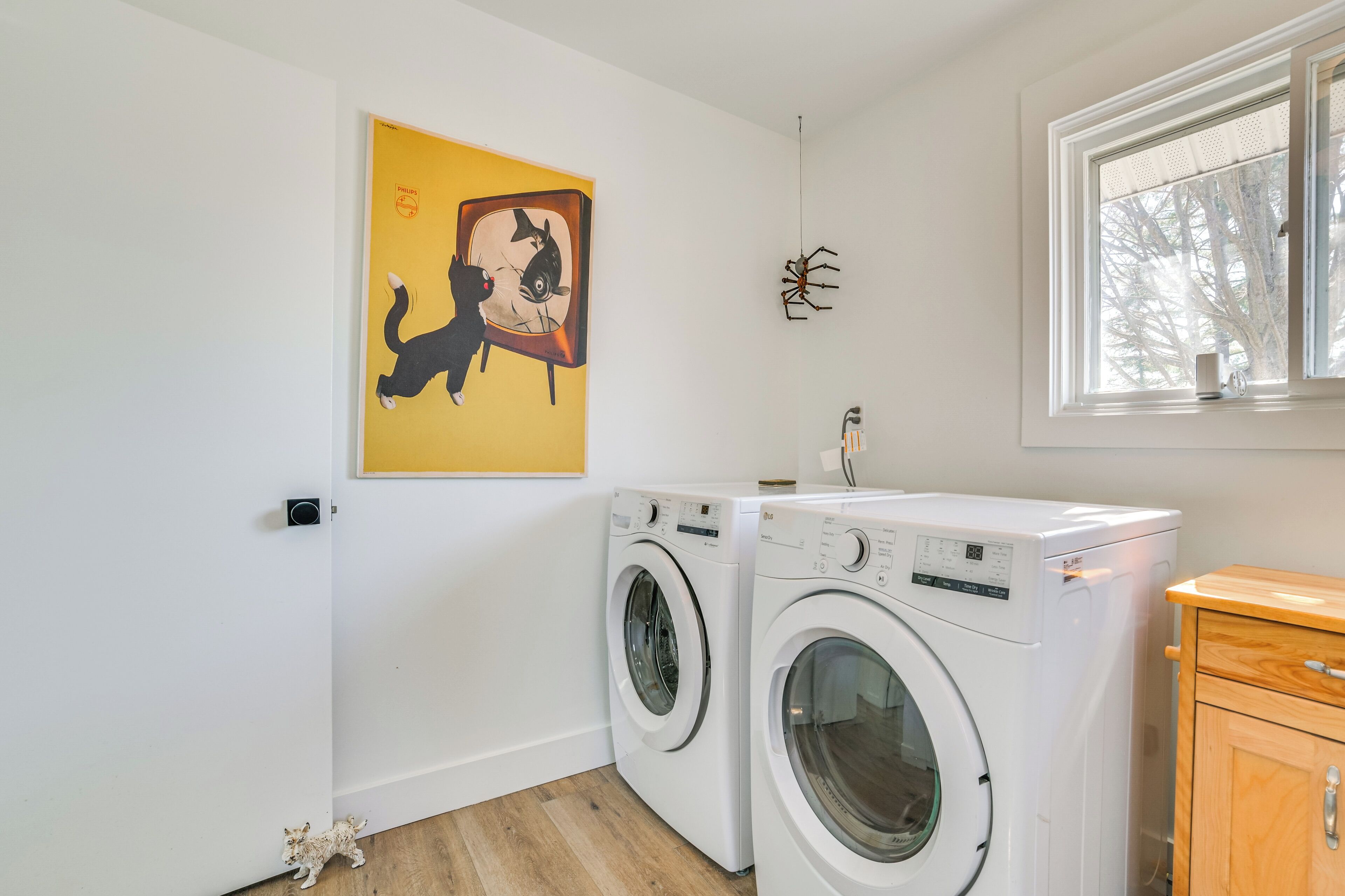 A laundry room with two white washing machines, a wooden cabinet, a yellow painting, and a spider decoration on the wall.