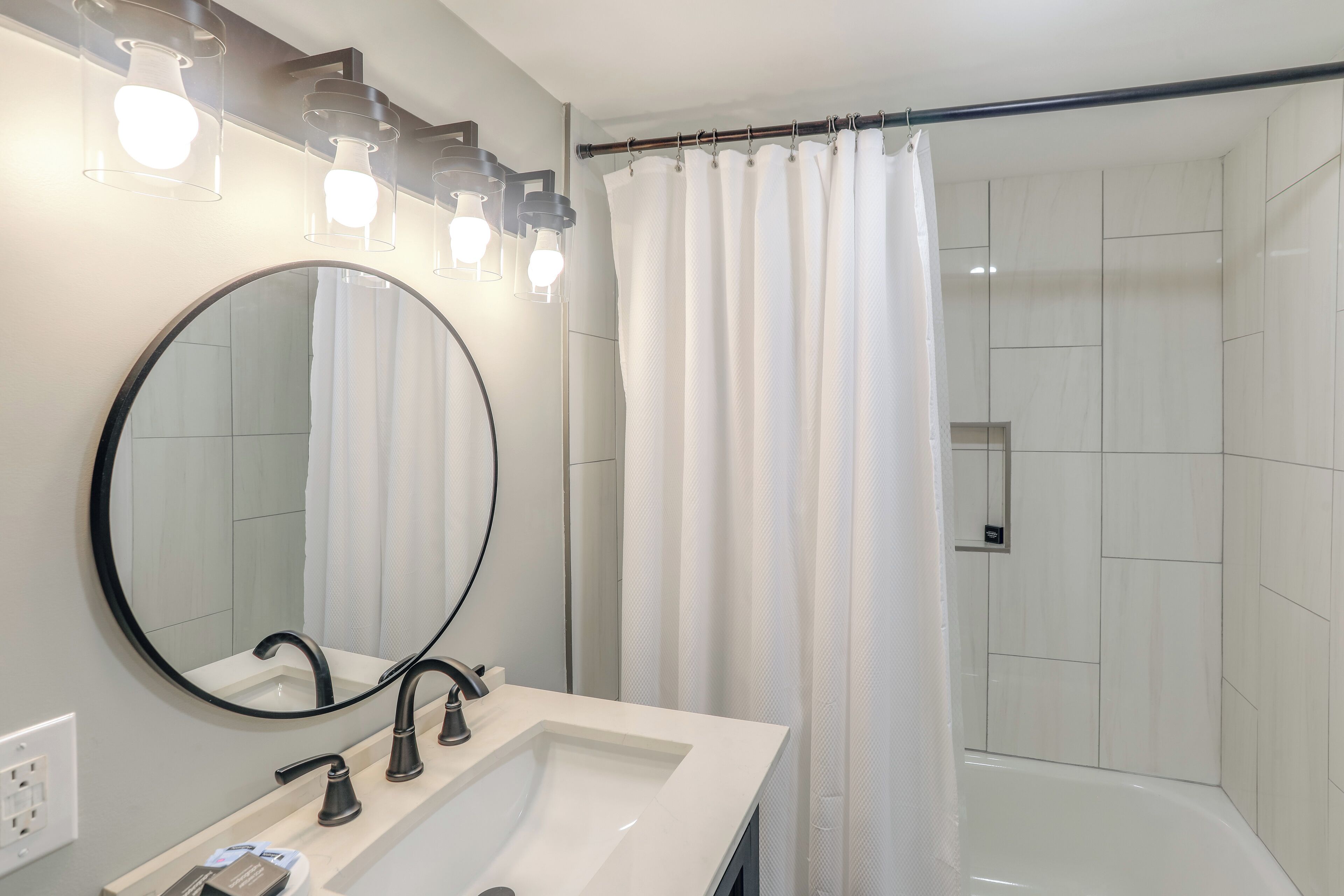 Une salle de bain moderne avec un lavabo blanc, un robinet noir et un grand miroir. On peut voir un rideau de douche et une baignoire.