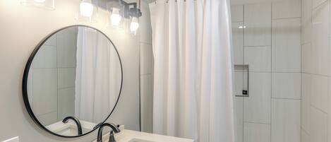 Une salle de bain moderne avec un lavabo blanc, un robinet noir et un grand miroir. On peut voir un rideau de douche et une baignoire.