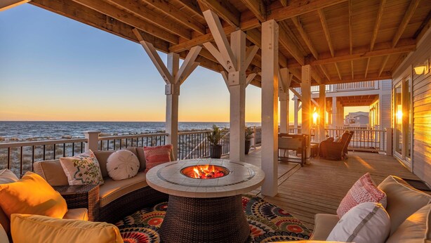 A wooden deck with a fire pit, wicker furniture, and a view of the ocean at sunset.
