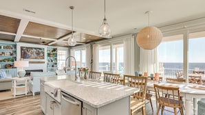 A spacious living area with a kitchen island, dining table, and chairs. There is a large window offering a view of the ocean.