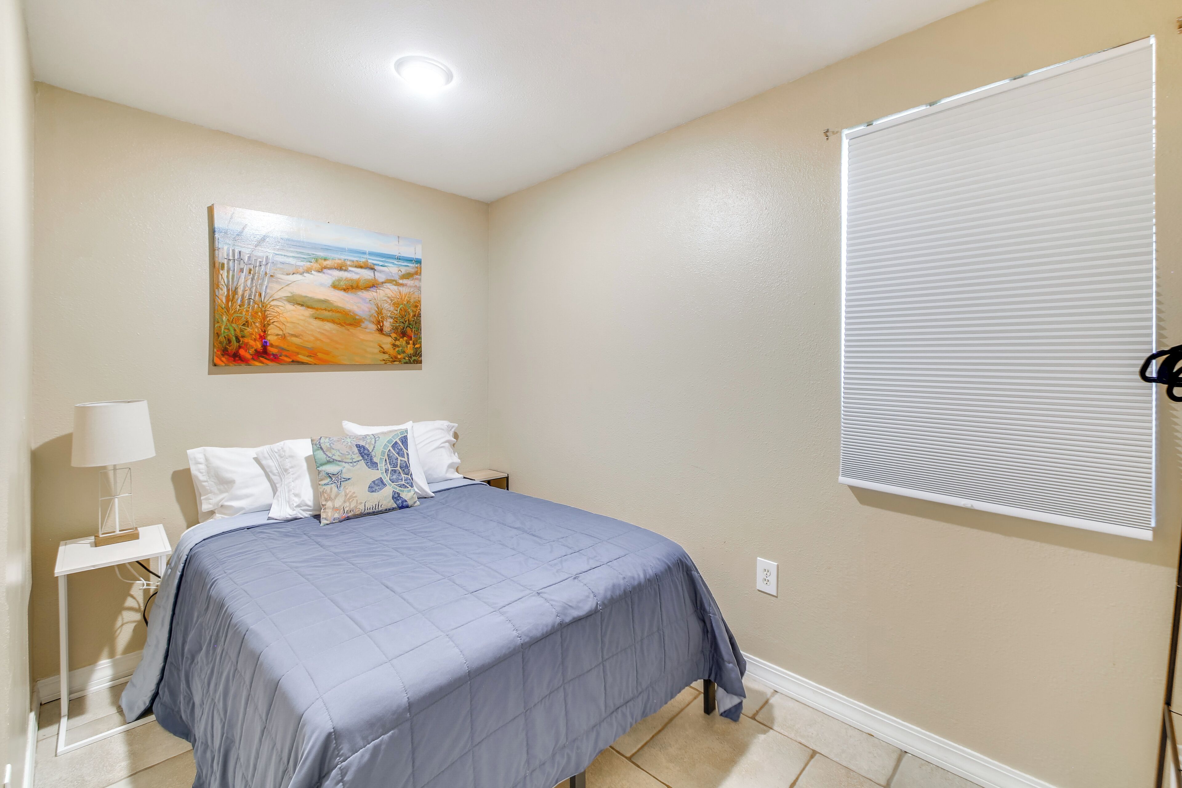 A bedroom with a bed, a nightstand, a lamp, a painting, and a window with blinds.