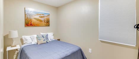 A bedroom with a bed, a nightstand, a lamp, a painting, and a window with blinds.