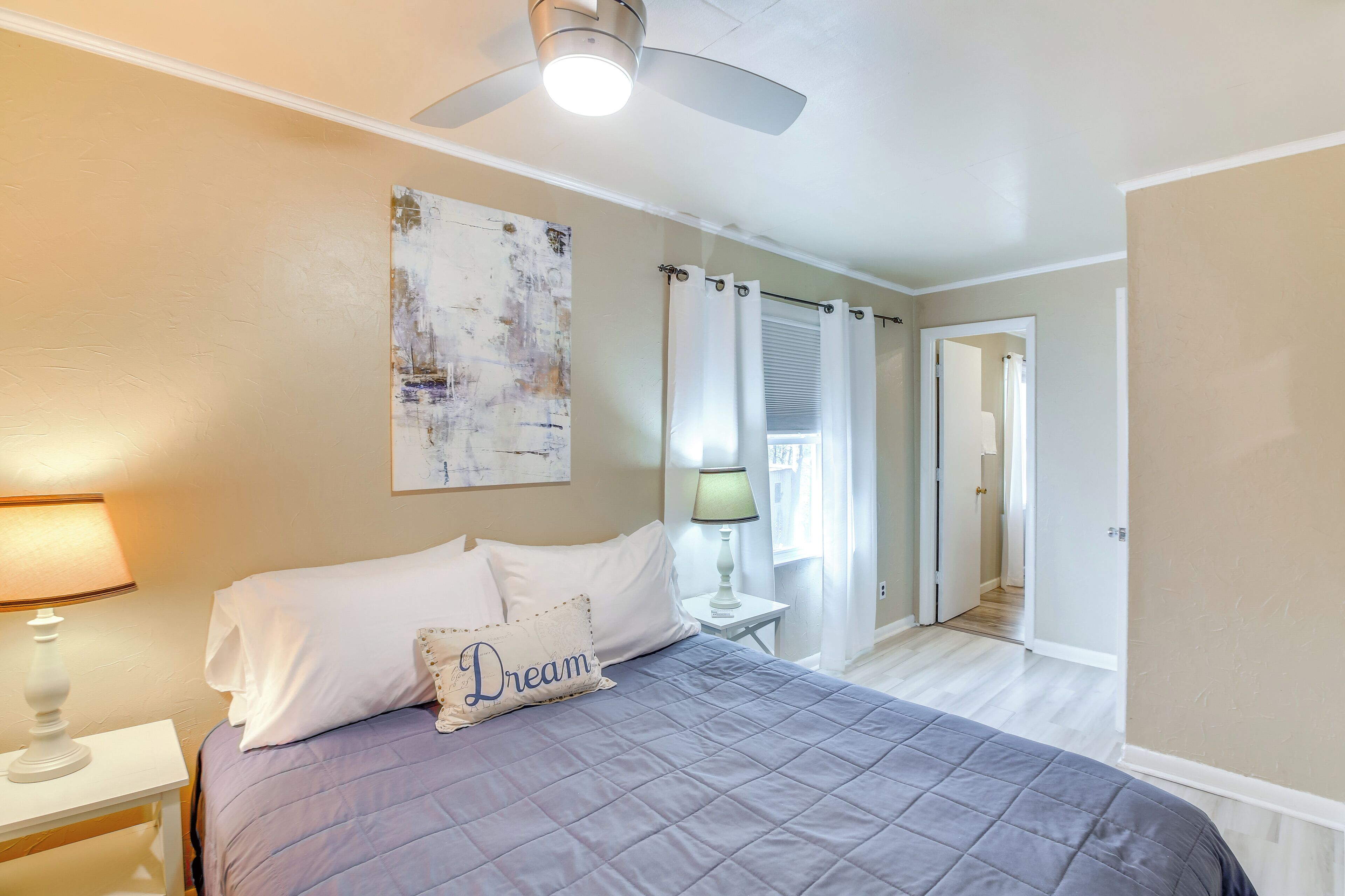 Une chambre à coucher avec un lit, un ventilateur de plafond, une table de chevet avec une lampe, un tableau au mur et une porte donnant sur une autre pièce.