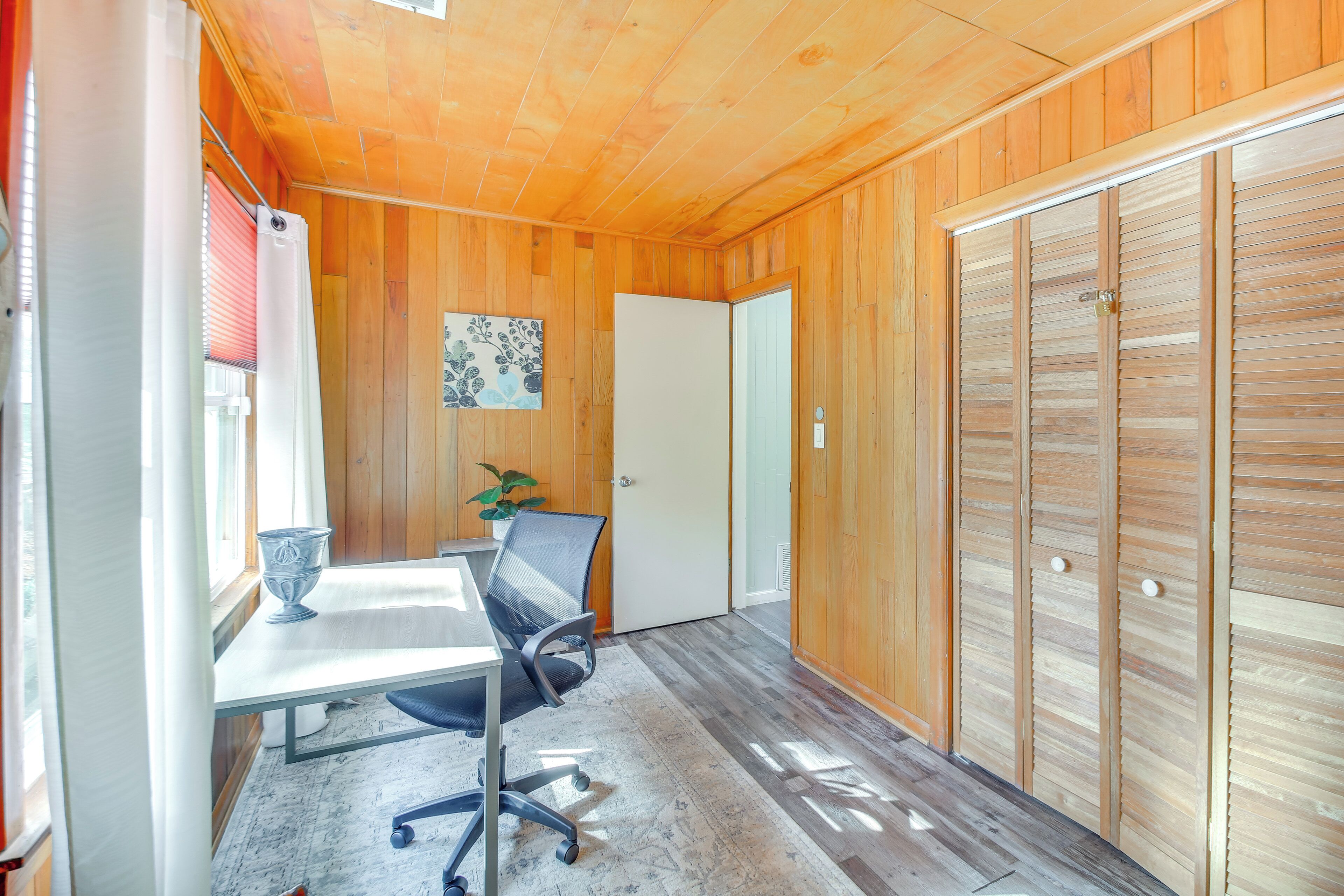 A room with wooden walls, a white door, a table with a chair, and a potted plant.