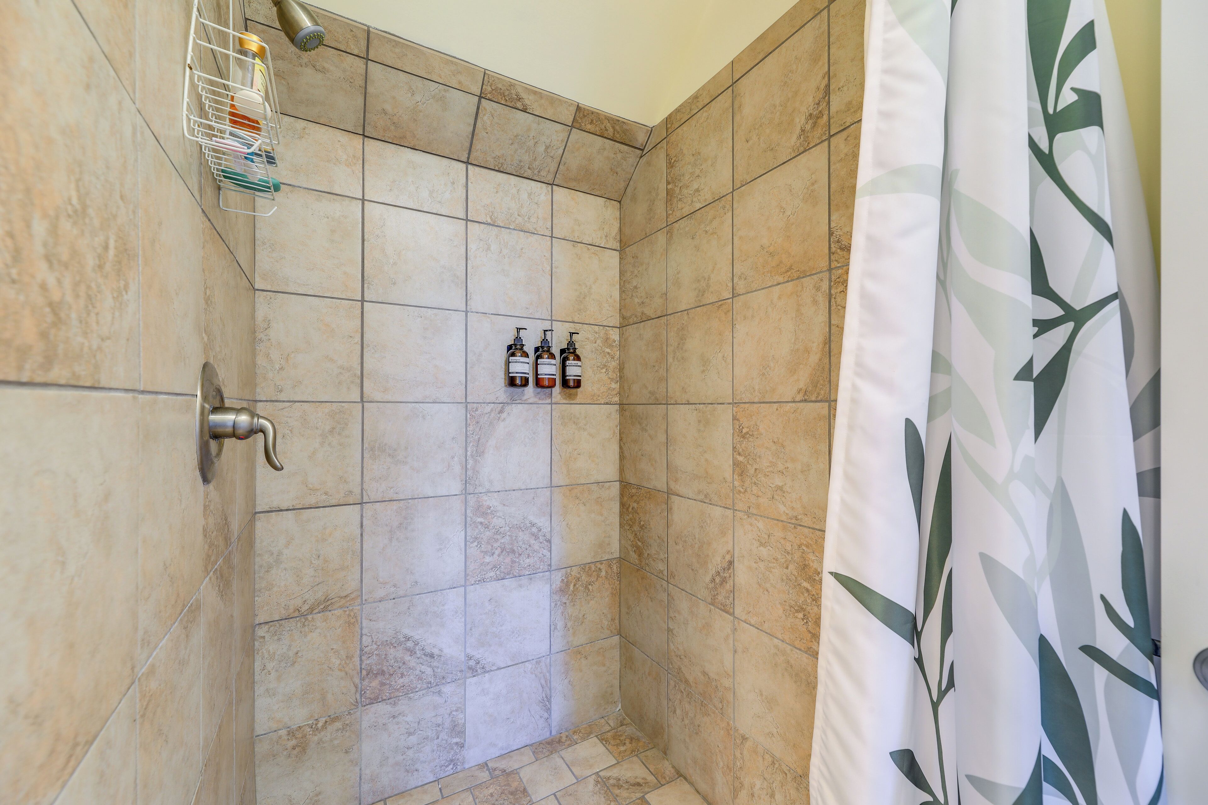 A shower with beige tiles, a metal showerhead, and two bottles of shampoo and conditioner on the wall.