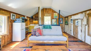 Ein gemütliches Blockhaus-Interieur mit Deckenventilator aus Holz, einer Couch mit bunten Kissen, einer Küchenzeile mit Kühlschrank und einem großen Fenster.