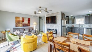 Ein modernes Wohnzimmer mit einer Couch, einem gelben Hocker und einem Esstisch mit Stühlen. Es gibt einen Kamin, einen Deckenventilator und eine Küchenzeile mit Schränken und Geräten.