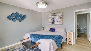 Ein Schlafzimmer mit Bett, Nachttischen, einem Sessel, Deckenventilator, Wandbildern und einer Tür zu einem anderen Raum.