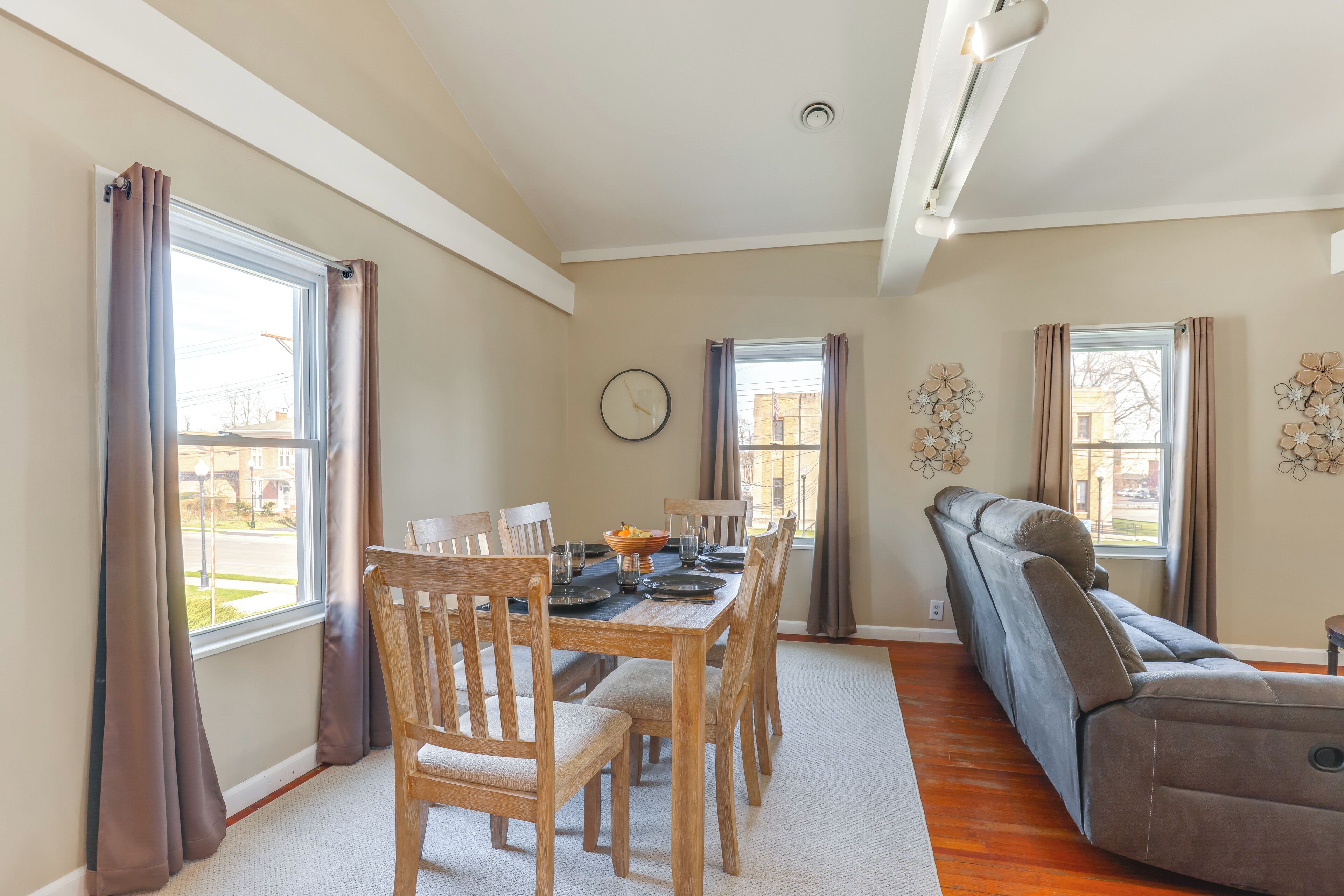 A dining area with a wooden table and chairs, a reclining sofa, and large windows with curtains.
