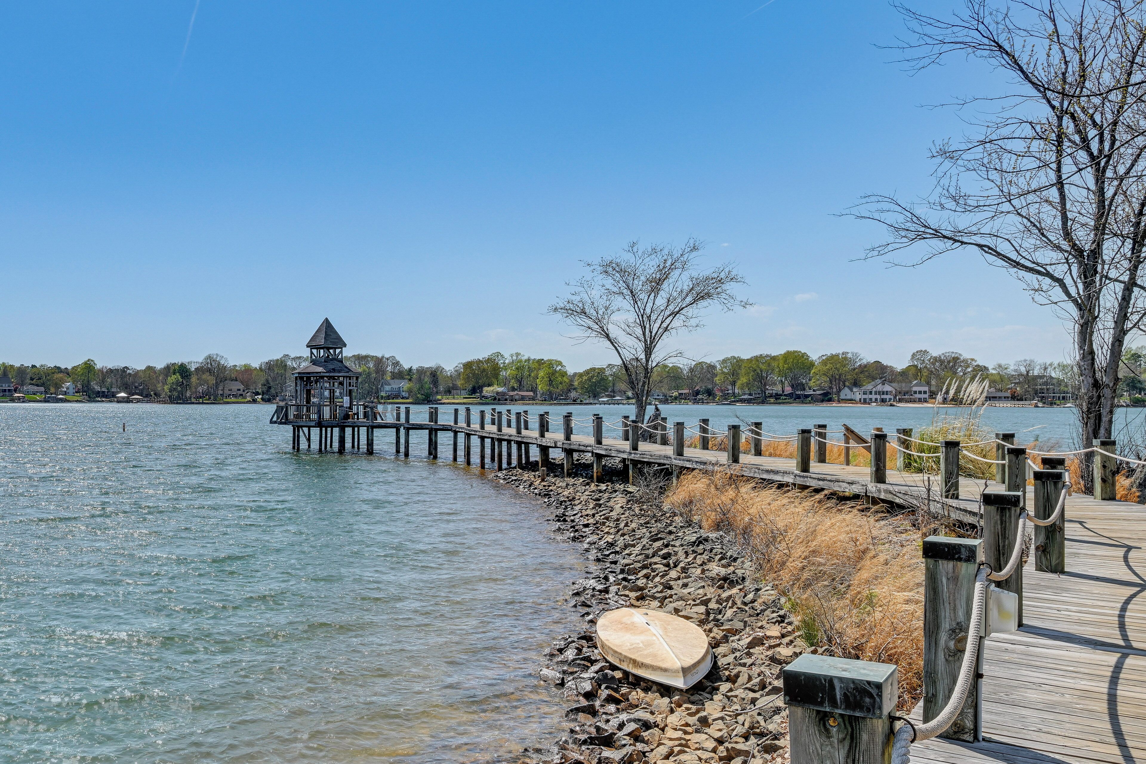 A wooden pier leading to a gazebo by a calm lake, with a boat on the shore and a clear sky.