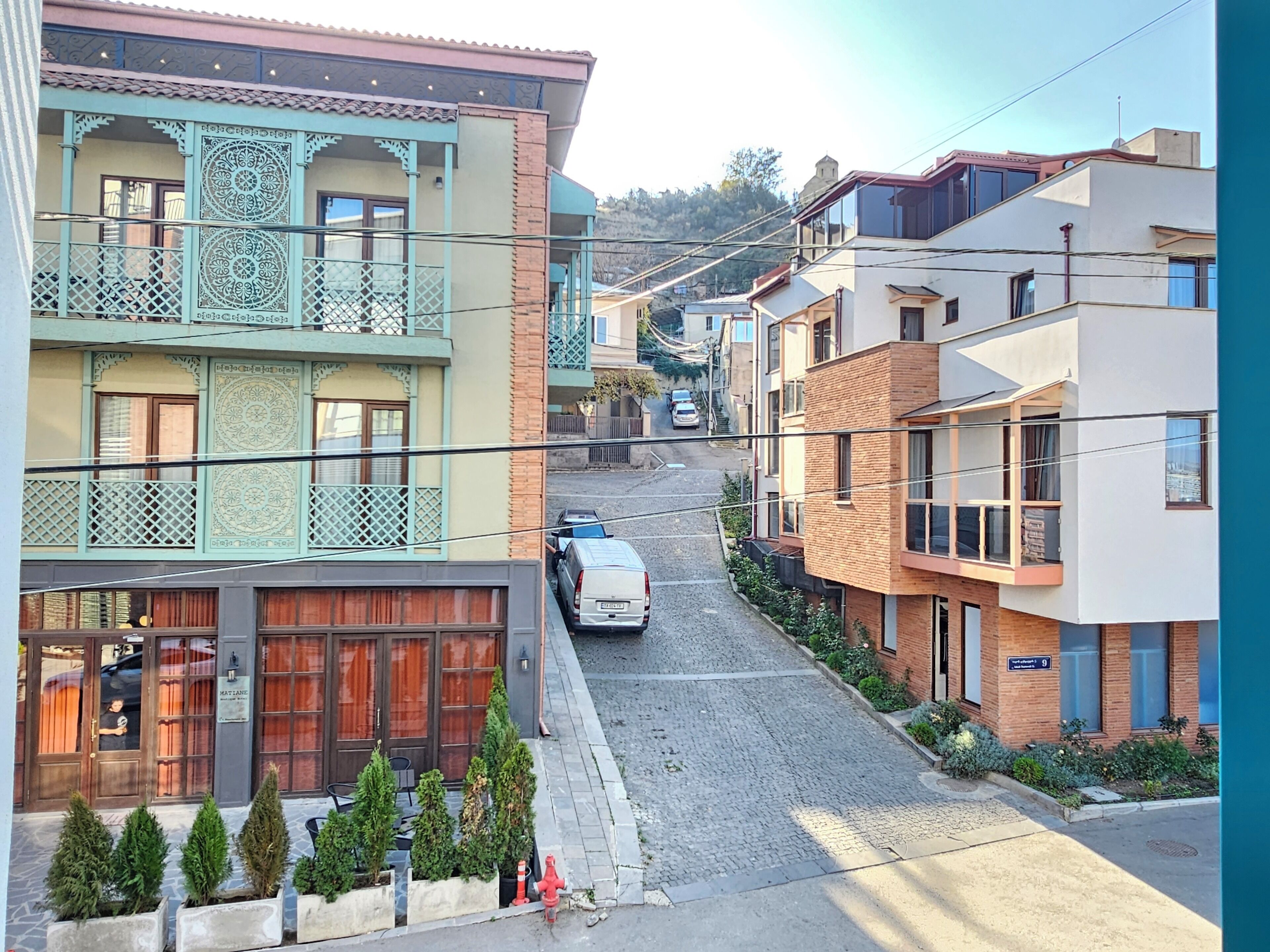 Une rue étroite bordée d’immeubles à plusieurs étages, dont certains sont dotés de balcons et de rampes décoratives. Un fourgon blanc est garé dans la rue, et quelques plantes en pot sont disposées le long du trottoir.