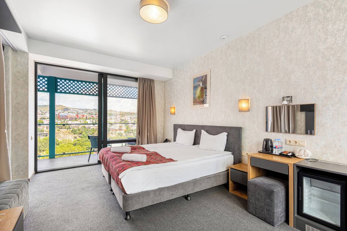 A hotel room with a bed, a desk, a television, and a balcony with a view.