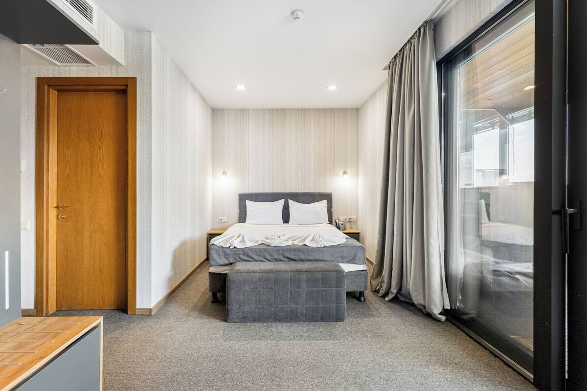 A modern hotel room with a large bed, a bedside table, a bench, a wardrobe, and a sliding glass door to the outside.