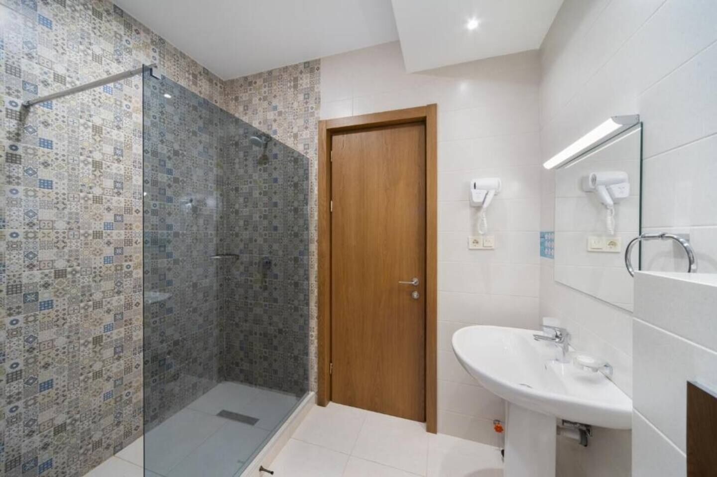 Un baño con cabina de ducha de vidrio, puerta de madera, lavamanos blanco y espejo con luz superior.