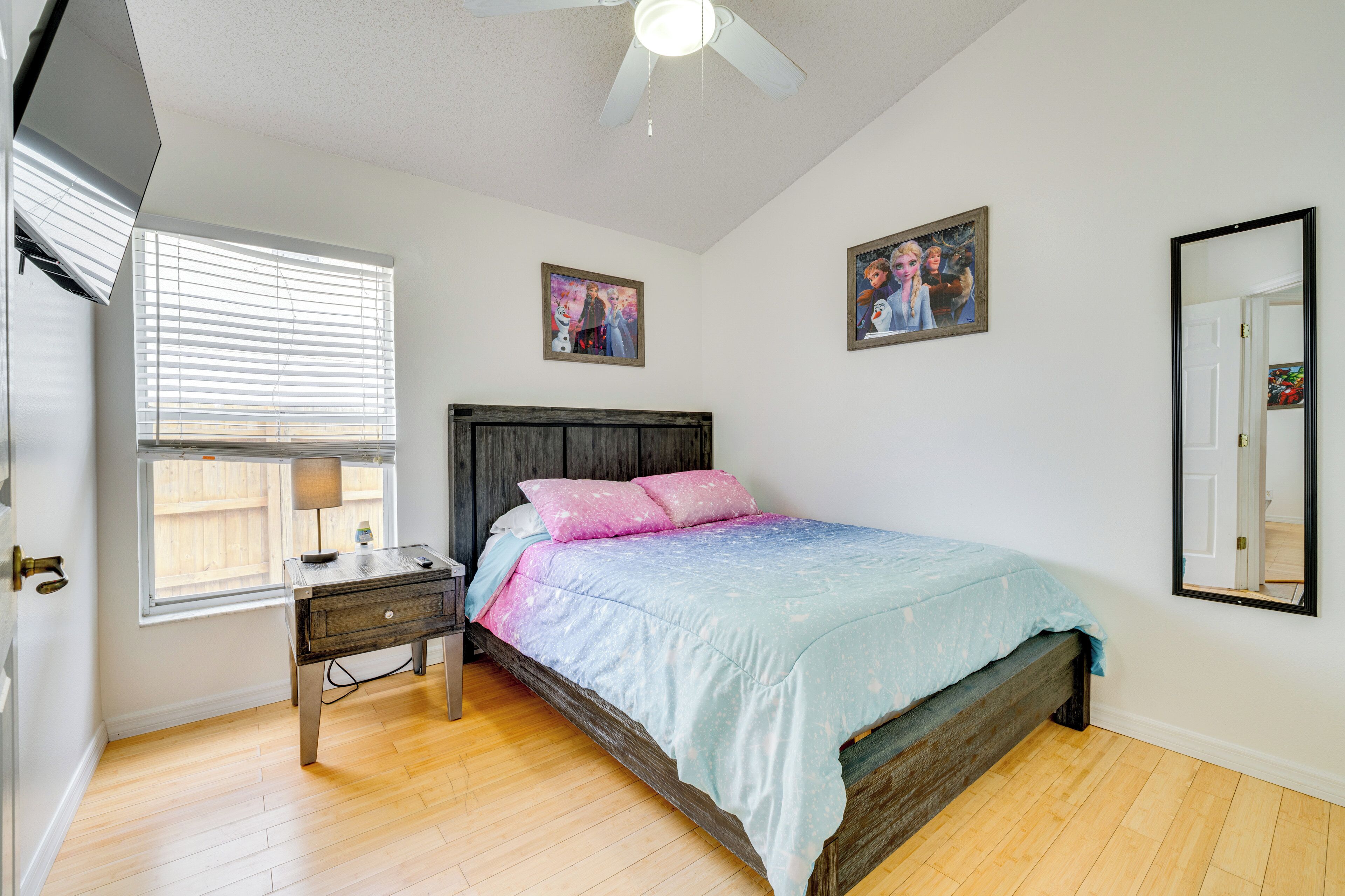 A bedroom with a wooden bed, a nightstand, a desk, a window with blinds, and two framed pictures on the wall.