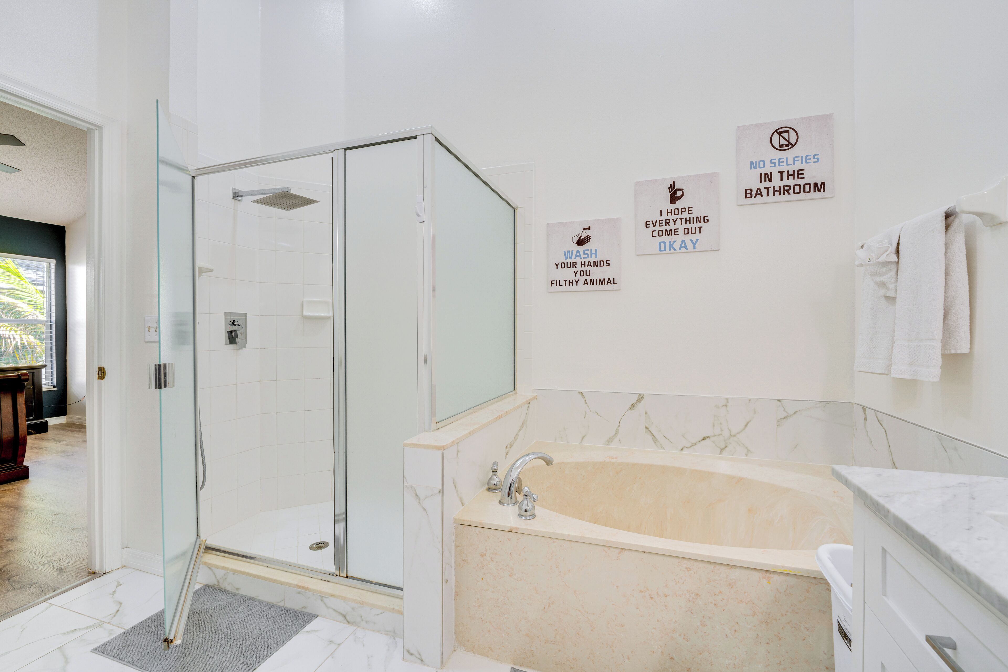 A modern bathroom with a glass-enclosed shower, a marble bathtub, and three wall signs with hygiene messages.