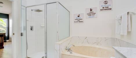 A modern bathroom with a glass-enclosed shower, a marble bathtub, and three wall signs with hygiene messages.