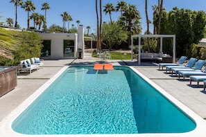 A rectangular pool with lounge chairs, a covered seating area, and a modern building with a glass door.