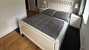 Une chambre à coucher avec un lit blanc, une table de chevet, une lampe et une fenêtre.