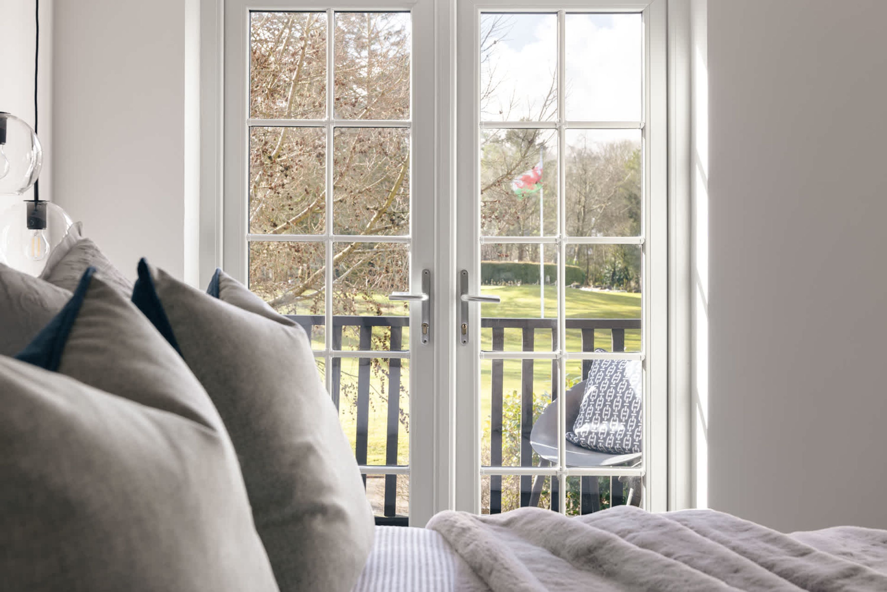A bedroom with a large window, a bed with pillows, and a view of a garden and deck.