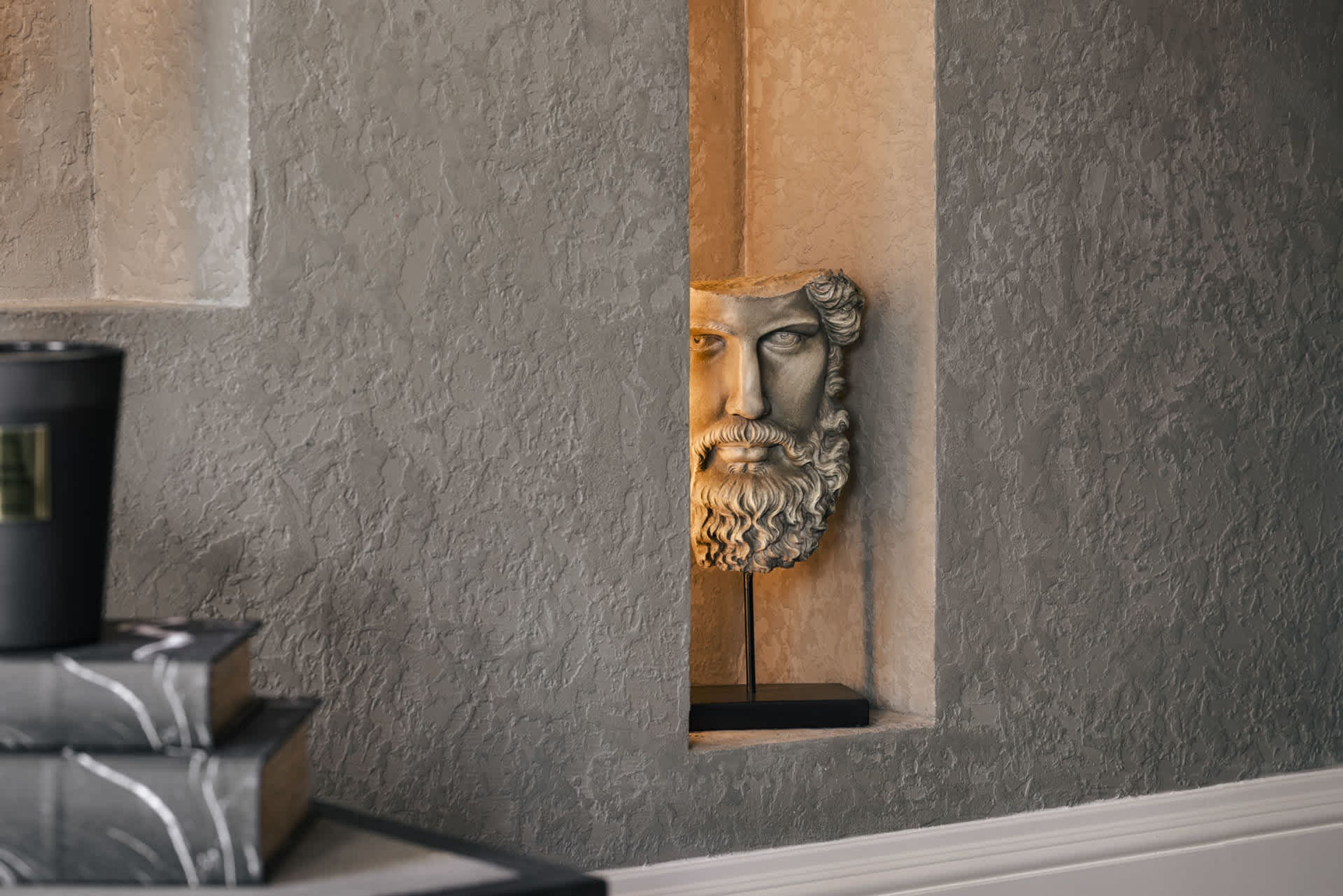Un mur de pierre orné d’un buste sculpté dans une niche, une pile de livres et un objet cylindrique noir.