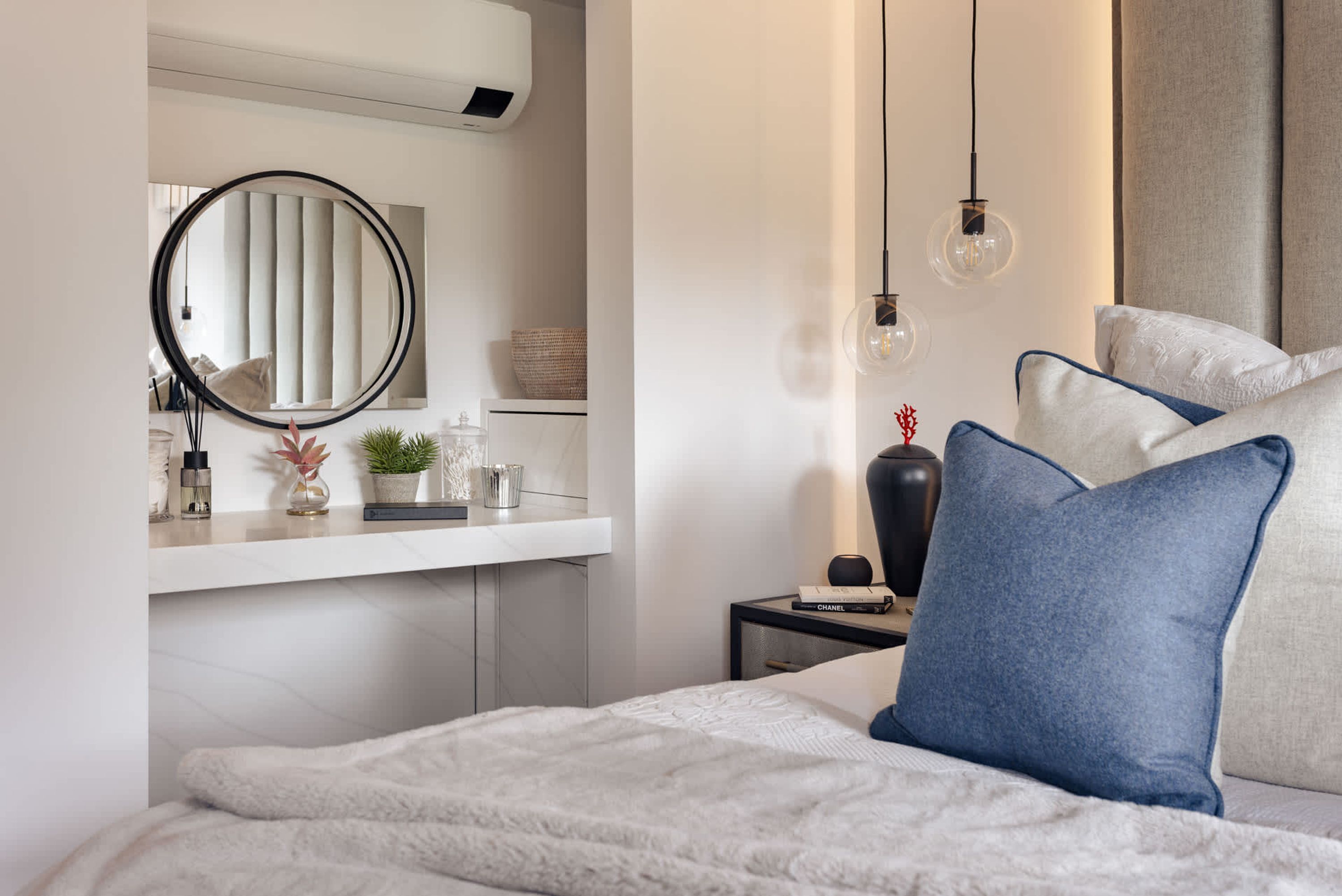 Een slaapkamer met een bed, kussens, een nachtkastje, een spiegel en een plank met decoratieve objecten.